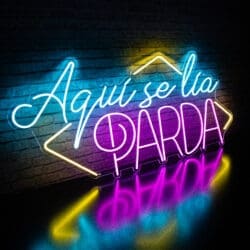 Alquiler Neón Aquí se Lía Parda con el texto en español "Aquí se lía parda", iluminado en azul, amarillo y rosa sobre un fondo de pared de ladrillos.