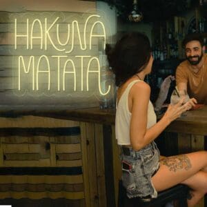 Una mujer sentada en un bar Neón Hakuna Matata con un hombre al fondo.