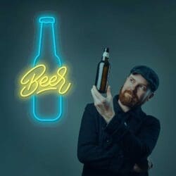Un hombre con gorra y chaqueta inspecciona una botella de cerveza Neón Botella, parado junto a un letrero de neón brillante que dice "Cerveza".