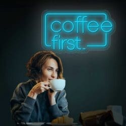 Una mujer disfrutando de una taza de Neón Coffe First debajo de un letrero de neón que dice "Coffe First" en un ambiente con poca luz.