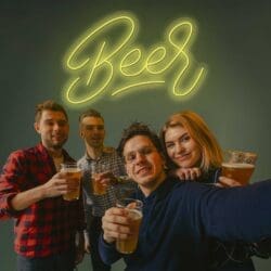 Cuatro amigos animan con vasos de cerveza Neón Letrero frente a un letrero de neón de "cerveza" con forma de botella, sonriendo hacia la cámara.