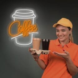 Una mujer con uniforme y gorra naranja sonriendo y presentando un soporte de cartón con dos tazas de café para llevar, con un letrero brillante de Neón Vaso de Café al fondo.