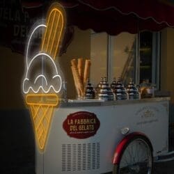 Un carrito callejero de helados con tres grandes contenedores bajo un cartel de neón de "Neón Helado Barquillo", junto al toldo de un restaurante.