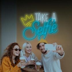Tres jóvenes adultos tomándose un selfie en la mesa de un café bajo un letrero de neón que dice "Neón Take a Selfie".