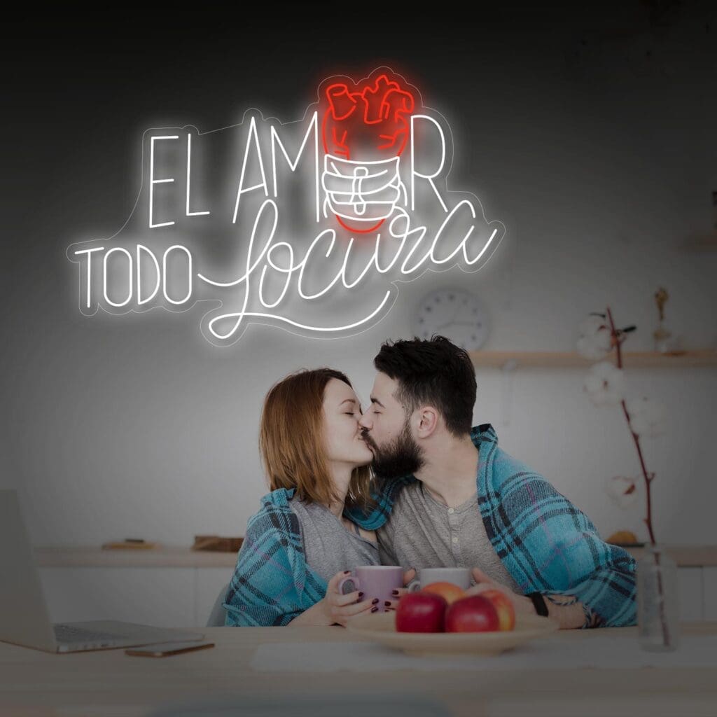 Una pareja sentada en una cocina, envuelta en mantas y sosteniendo tazas, mientras se besa. Un letrero de neón detrás de ellos dice "Neón El Amor Todo Locura" con un gráfico de corazón y cerebro. Sobre la mesa hay un cuenco de manzanas que le da un toque acogedor. El mensaje iluminado les incita a tomarse una selfie mientras saborean el momento.