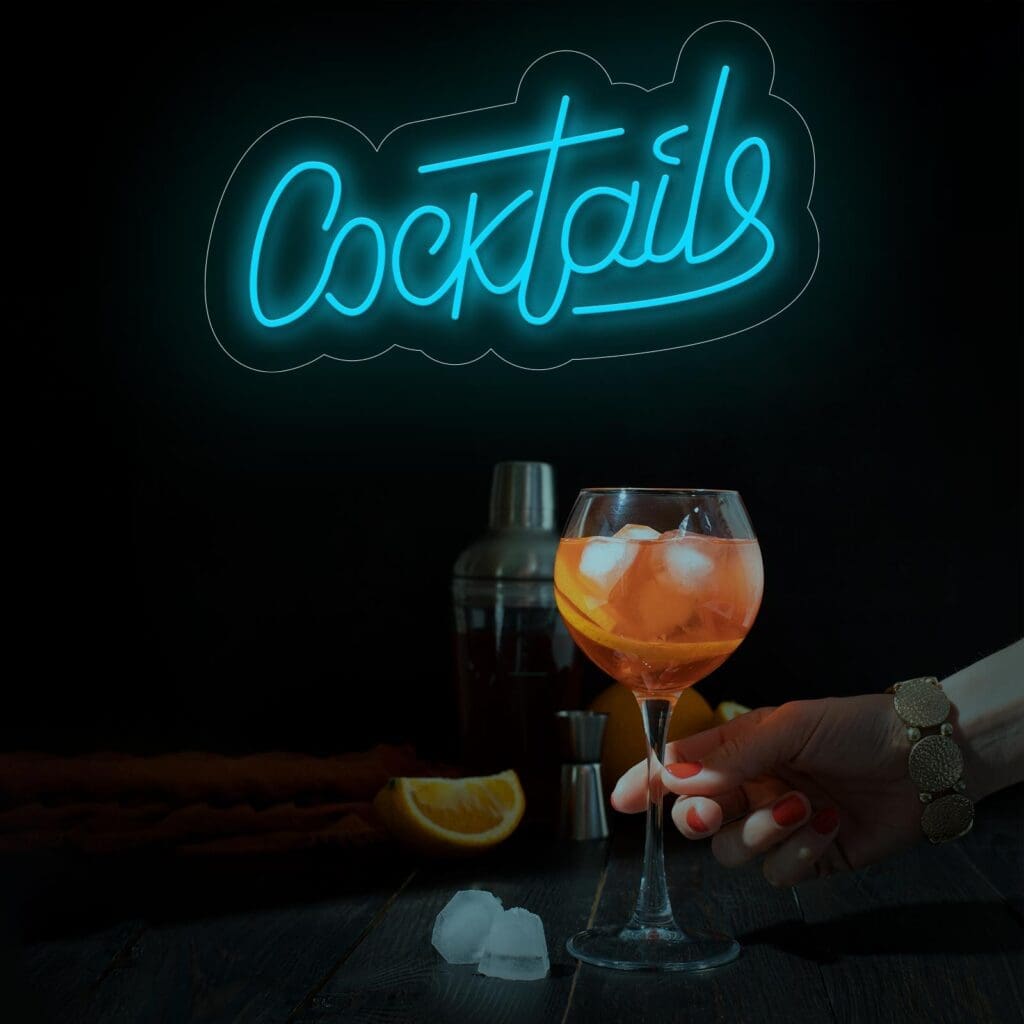 Una mano sosteniendo una copa de cóctel con bebida y hielo, frente a un letrero de neón que dice "Neón Letrero COCKTAILS". Hay cubitos de hielo y rodajas de naranja sobre la mesa. Al fondo también se ve una coctelera. ¡Lugar perfecto para tomarse una selfie!