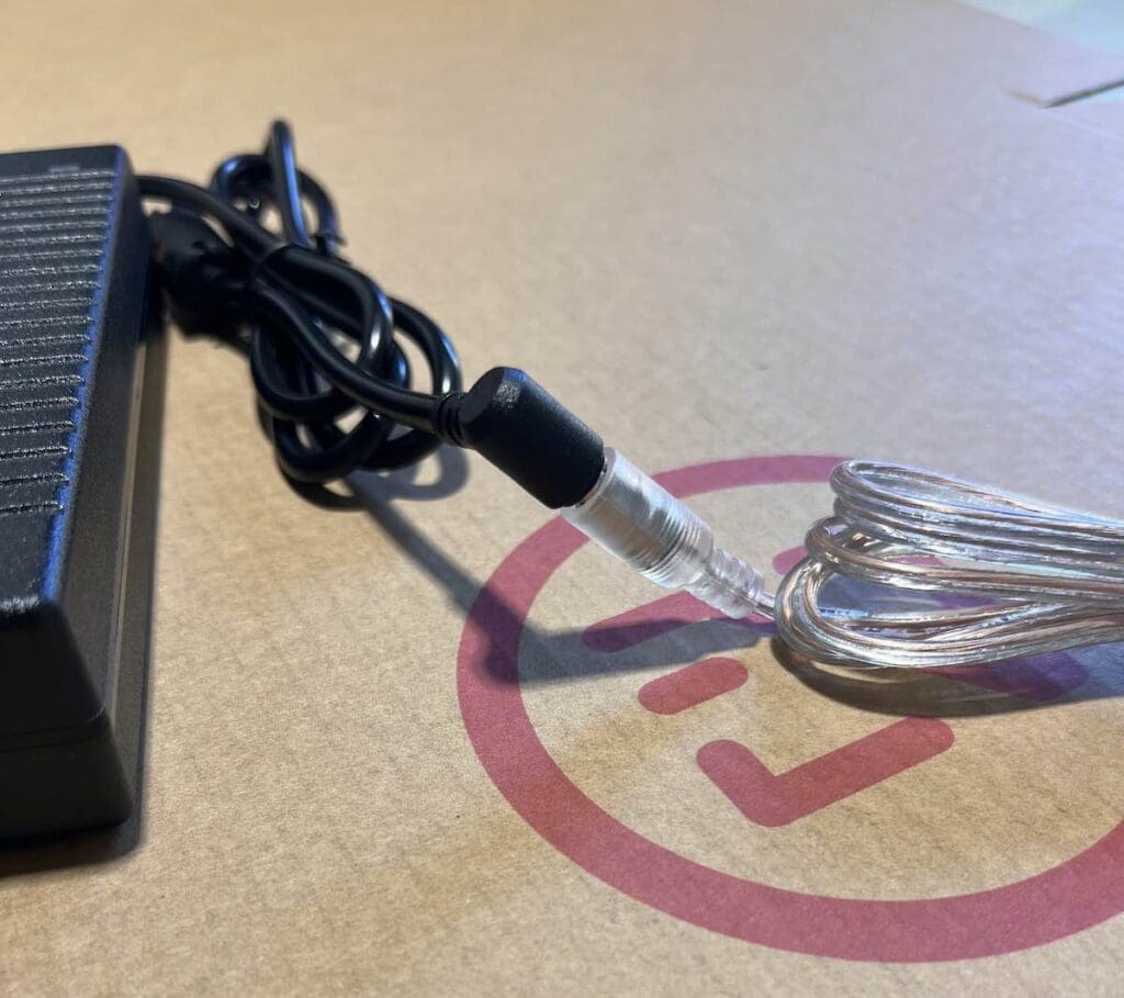 Primer plano de un adaptador de corriente negro conectado a un cable plateado enrollado con una funda transparente, colocado sobre una superficie de cartón con un logotipo circular rojo.