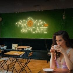 Una mujer bebe de una taza blanca en un café con mesas y sillas de madera. Un letrero de neón en la pared verde del fondo dice "Aroma de Café".