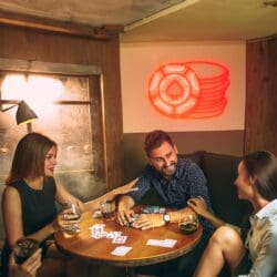 Tres personas están sentadas en una mesa redonda en una habitación con poca luz, jugando a las cartas y bebiendo. Hay un letrero de neón con el símbolo de una pica en la pared detrás de ellas.