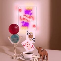 Tarjeta del Rey de Corazones iluminada con neón en la pared, vaso con esferas rojas y azules, naipes, taza de café, figura de gato de madera y dominó en la mesa.