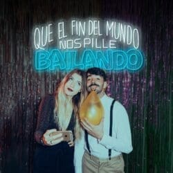 Una mujer y un hombre sonriendo y posando para una foto en un entorno festivo con un texto de neón en español encima de ellos que dice: "Que el fin del mundo nos pille bailando".