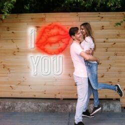 Una pareja se abraza felizmente frente a una pared de madera con un letrero de neón que muestra una marca de beso y las palabras "YO [marca de beso] TÚ".