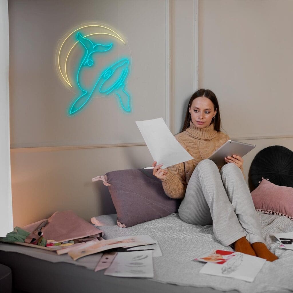 Una mujer está sentada en una cama revisando papeles con una tableta en la mano. Una ballena de neón y la luz de la luna decoran la pared detrás de ella. A su alrededor hay varios materiales de arte y dibujos.