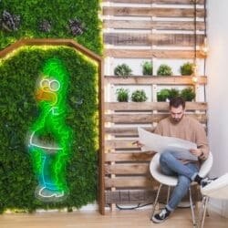 Un hombre se sienta en una silla leyendo un periódico grande en una habitación con una pared de madera, plantas y un contorno de neón de un personaje de dibujos animados en un panel vertical de plantas verdes.