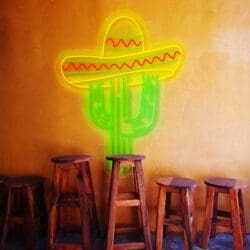 Letrero de neón con forma de cactus y sombrero sobre una pared amarilla encima de cinco taburetes de bar de madera.