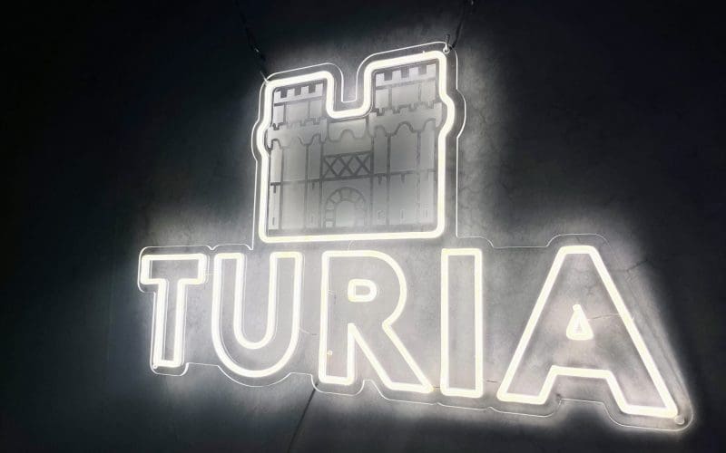 Letrero de neón iluminado que dice "TURIA" debajo del ícono de un castillo. El cartel brilla sobre un fondo oscuro.